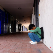 A distressed young adult sitting alone in a school hallway, symbolizing rising suicide, behavioral health challenges, and preventable deaths among young adults in the United States.