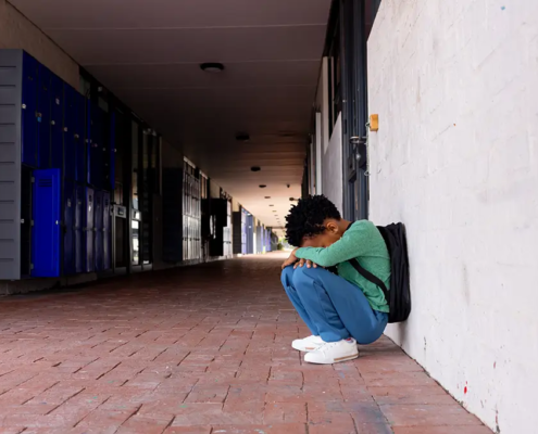 A distressed young adult sitting alone in a school hallway, symbolizing rising suicide, behavioral health challenges, and preventable deaths among young adults in the United States.
