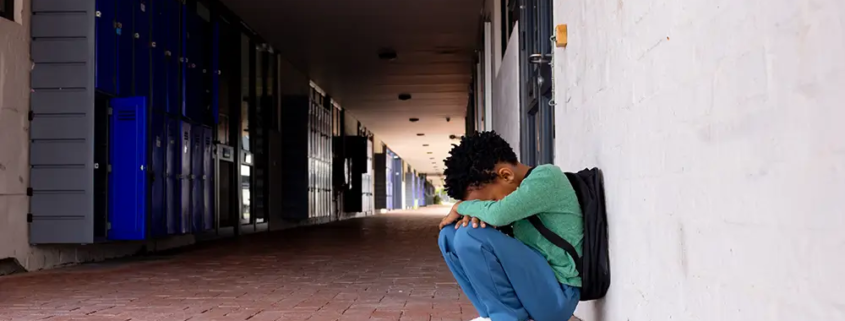 A distressed young adult sitting alone in a school hallway, symbolizing rising suicide, behavioral health challenges, and preventable deaths among young adults in the United States.